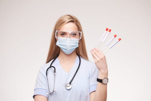 A female healthcare professional wearing a mask and protective goggles, holding test tubes with red caps. She has long hair and a stethoscope around her neck, standing against a neutral background.