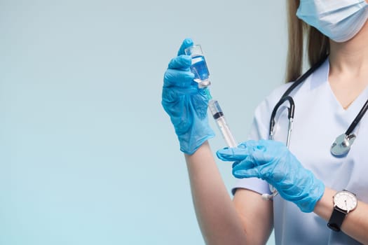 A healthcare professional in scrubs and gloves prepares a syringe with a blue liquid. They are wearing a mask and have a stethoscope around their neck, set against a light blue background.