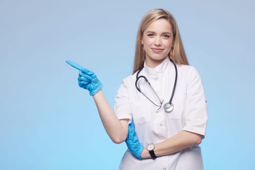 Beautiful lady doctor pointing with hand to copy space, studio shot. Isolated on blue background. Medical student, general practitioner.