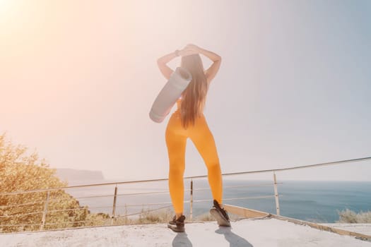 A woman wearing yellow leggings and a sports bra is standing on a cliff, looking out at the ocean with a rolled up yoga mat on her shoulder.