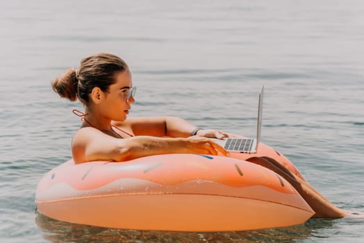 Woman freelancer works on laptop swimming in sea on pink inflatable ring. Pretty lady typing on computer while floating in the sea on inflatable donut at sunset. Freelance, remote work on vacation