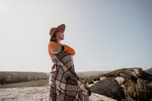 A woman wearing a hat, a warm sweater, and a plaid blanket is standing on a hill, looking out at the landscape in front of her.