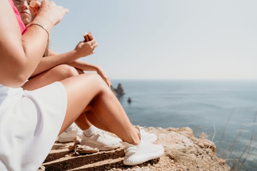 Two women wearing white skirts and sneakers sit on a cliff overlooking the ocean on a sunny day.