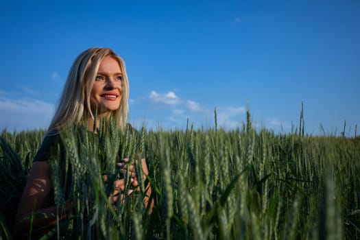 On a sunny day, a young and attractive female farmer stands proudly in her wheat field, overseeing her bountiful crops. Dressed in practical work attire, her blonde hair catching the sunlight, she exudes a sense of vitality and dedication. With a warm smile revealing her pearly white teeth, she radiates contentment and satisfaction. The wheat stalks tower around her, almost reaching her height, their vibrant green hue a striking contrast against the blue sky. As she surveys the golden expanse before her, her satisfaction with the abundant harvest is evident. Against the backdrop of the sunlit landscape, her presence adds a touch of rural charm to the scene. She looks ahead with smile. This captivating image captures the essence of agricultural life, where hard work and perseverance yield fruitful rewards, and the beauty of nature is celebrated with every successful harvest