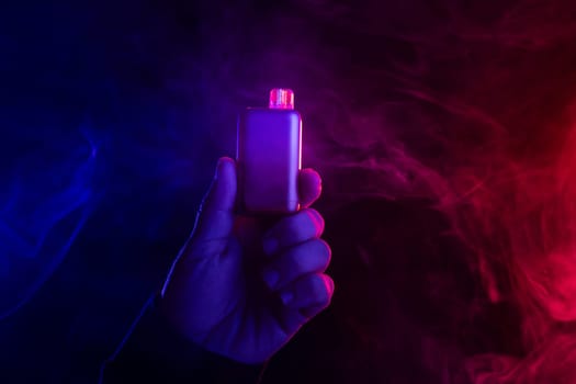 close-up hand holding vape electronic cigarette against colored smoke background, concept of harm of smoking