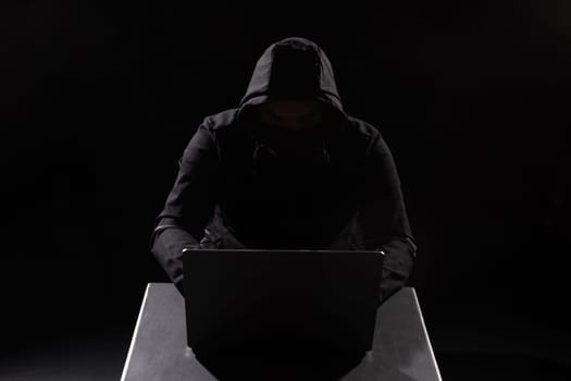 Cyber hacker in hood behind laptop in dark room, digital cyber security concept, computer hacking, information security