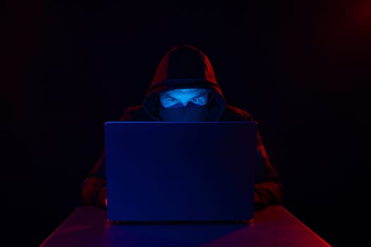 Cyber hacker in hood behind laptop on dark background with red blue light, anonymous spy. digital cyber security concept