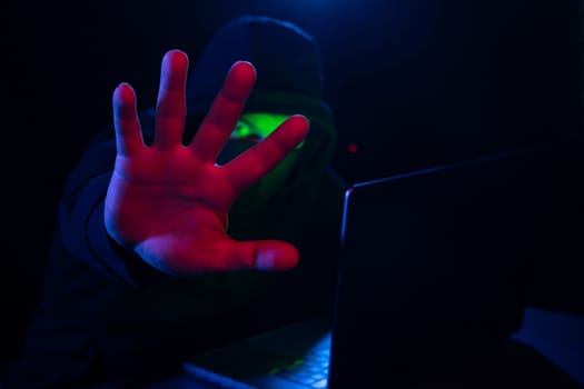 Cyber hacker in hood behind laptop puts hand forward hiding his face in dark room, concept of digital information protection, Computer hacking, stop cyber hacker