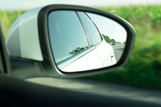 Rear-view mirror, close-up, with the reflection of a white car when the car is moving against the background of green trees. Soft focus. An essential element for driving safety.