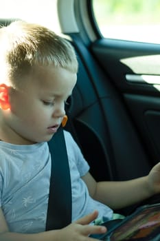 Security concept. A 4-year-old boy is riding in a car with a seat belt fastened in a child seat in the back seat. He travels with his family and watches cartoons on his iPad. Soft focus. vertical