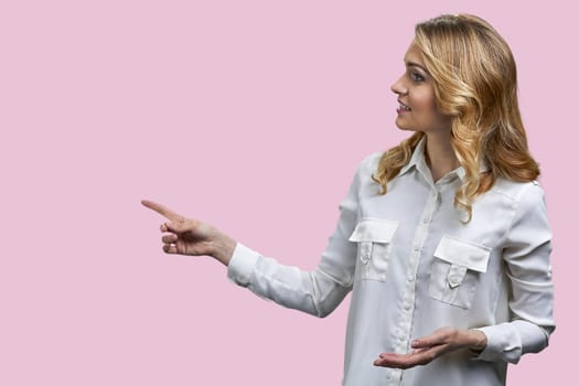 Attractive young blonde woman pointing at copy space at the left with her finger. Isolated on pink background.