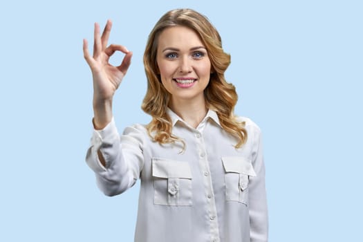 Portrait of a young blond smiling woman showing okay gesture. Cute lady isolated on blue.