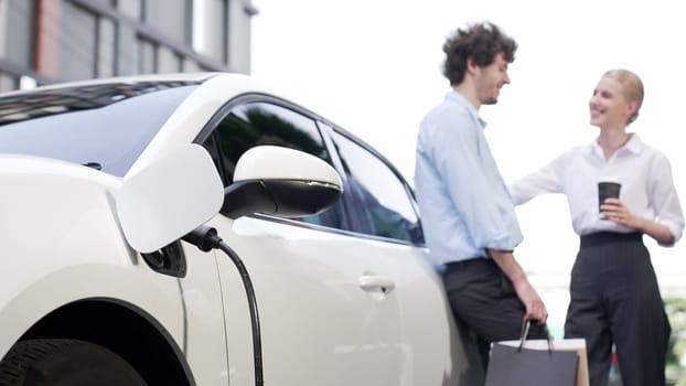 Progressive businessman and businesswoman leaning on electric car connected to charging station before driving around city center. Eco friendly rechargeable car powered by clean energy.