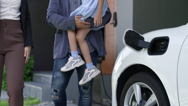 Progressive young parents and daughter living in a home with an electric vehicle and electronic vehicle charging station. Green and clean energy from electric vehicles for healthy environment.