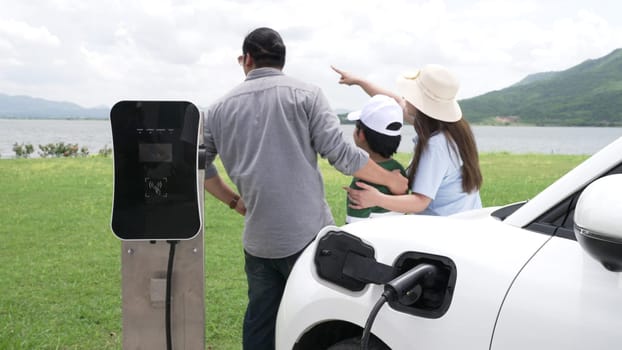 Concept of progressive happy family enjoying their time at wind farm with electric vehicle. Electric vehicle driven by clean renewable energy from wind turbine generator for charging station.