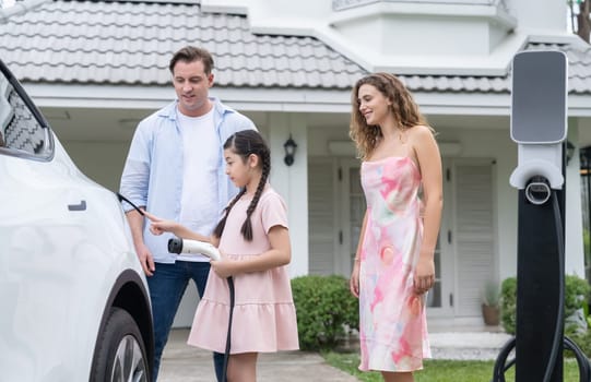 Happy little young girl learn about eco-friendly and energy sustainability as she help her family recharge electric vehicle from home EV charging station. EV car and modern family concept. Synchronos