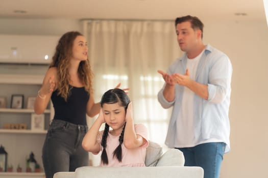 Annoyed and unhappy young girl sitting on sofa trapped in middle of tension by her parent argument in living room. Unhealthy domestic lifestyle and traumatic childhood develop to depression.Synchronos