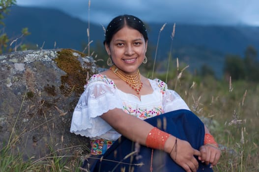 beautiful indigenous woman in traditional dress from otavalo, ecuador looking at the camera and smiling while sitting and leaning on a rock on the top of a mountain.Hispanic Heritage Month. High quality photo