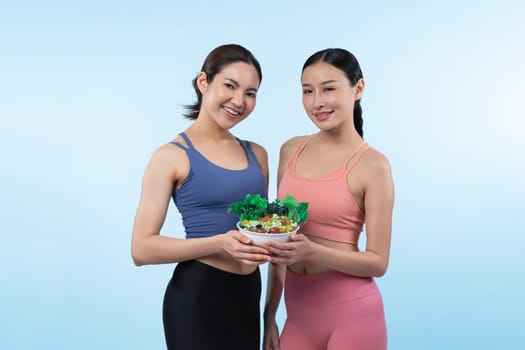 Two young sporty Asian women in sportswear holding salad bowl fill with fruit and vegetable. Natural youthful and fit body lifestyle people with balance nutrition on isolated background. Vigorous