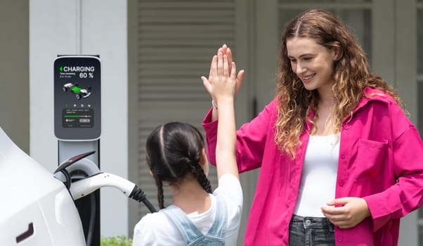 Happy little young girl learn about eco-friendly and energy sustainability as she help her mother recharge electric vehicle from home EV charging station. EV car and modern family. Panorama Synchronos