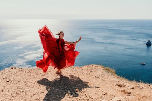 Side view a Young beautiful sensual woman in a red long dress posing on a rock high above the sea during sunrise. Girl on the nature on blue sky background. Fashion photo.