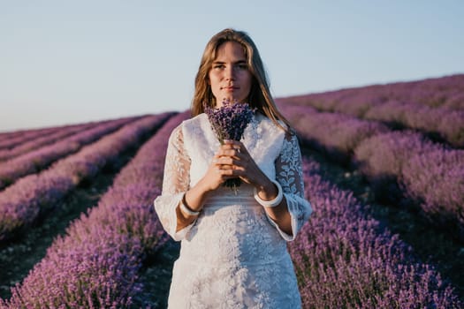 Close up portrait of young beautiful woman in a white dress and a hat is walking in the lavender field and smelling lavender bouquet.