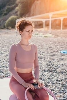 Middle aged well looking woman with black hair doing Pilates with the ring on the yoga mat near the sea on the pebble beach. Female fitness yoga concept. Healthy lifestyle, harmony and meditation.