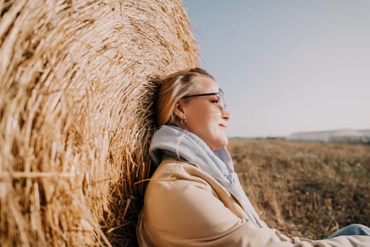 A woman in a beige jacket and blue hoodie leans against a hay bale, looking thoughtfully towards the horizon. The sky is bright blue and the field is a warm brown, creating a peaceful autumn scene.