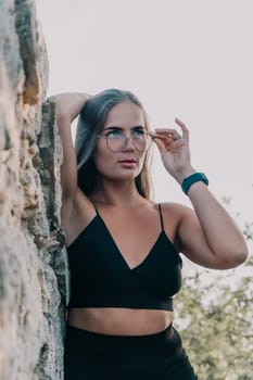 A woman wearing a black crop top and glasses leans against a rock, looking to her right.