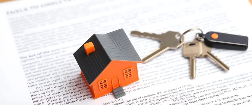 A small orange house model sits beside keys on a pile of printed documents related to real estate, highlighting the process of purchasing a new home.
