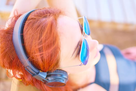 Fine woman listening to music on the beach. Redhead girl listening melody with headphones and smartphone while relaxing on lounger near ocean.