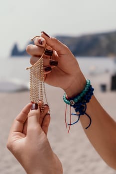 Two hands hold a gold chain necklace with multiple strands of beads, against a blurred beach background.