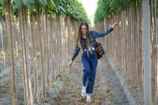 A cheerful young woman dressed in trendy and fashionable attire dances joyfully and freely amidst a backdrop of vibrant green trees in a serene, beautiful environment that evokes happiness