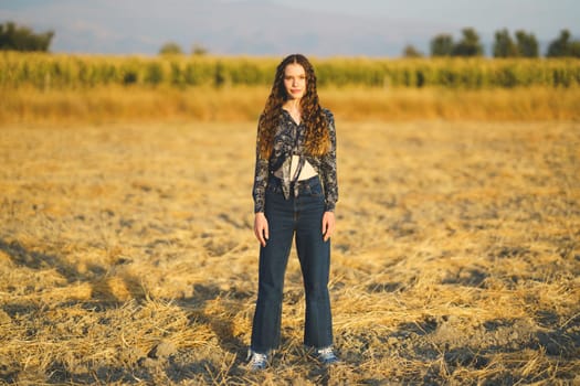 A young woman with long, curly hair stands proudly in a dry, golden field. The serene landscape features distant trees under a clear blue sky, creating a breathtaking and inviting scene