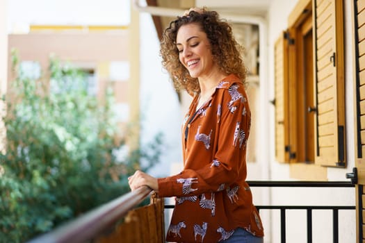 Side view of young female in stylish outfit smiling and looking down while standing on balcony against blurred background