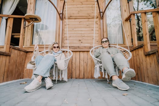 Two women wearing sunglasses and casual clothing relax in hanging chairs on a wooden porch.