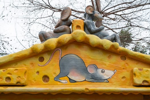 At a whimsical park, a colorful sculpture features three playful mice atop a yellow cheese structure. The scene captures the charm of autumn with bare trees in the background.