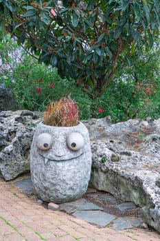 A decorative stone planter shaped like a smiling face sits in a garden, covered with grass as its hair and surrounded by vibrant greenery and colorful flowers.