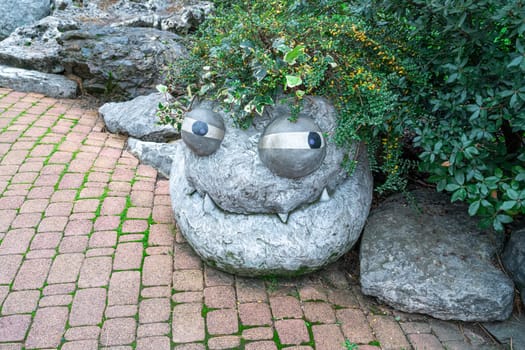 A large, rocky formation resembles a playful face, complete with eyes and teeth, adorned by lush greenery. It sits along a cobblestone pathway in a serene garden area.