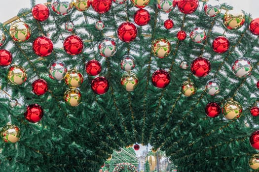 A beautifully adorned archway features vibrant red and gold ornaments amidst lush greenery, inviting visitors to explore a lively winter market filled with holiday spirit.