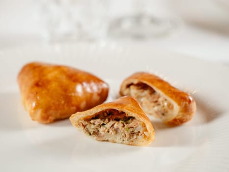 Small russian pies with minced tuna fish Russian Piroshki. Cooked tuna fish hand portioned pies or empanadas cut in half on white plate in elegant restaurant interior. Selective focus, close up.