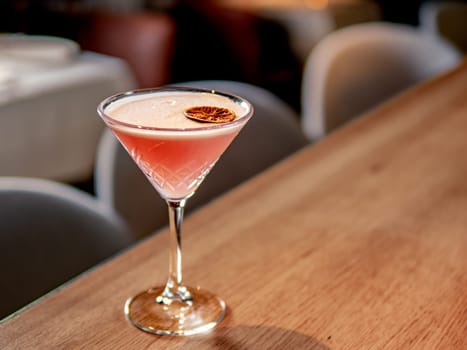 Red cocktail in martini glass on bar counter in restaurant interior. Gin alcohol cocktail decorated egg white and dry lichee fruit slice. Pink cold alcoholic cocktail in bar or nightclub background