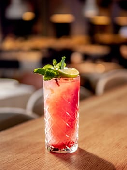 Red non-alcohol cocktail with crushed ice in glass on wooden bar counter in restaurant interior. Gin alcohol cocktail decorated lime and mint. Pink cold alcoholic cocktail in bar or nightclub background. Copy space
