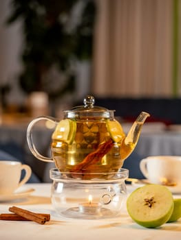 Herbal tea with apple and cinnamon in transparent glass teapot with warmer and candle on table in restaurant. Herbal tea concept, wellbeing and health care