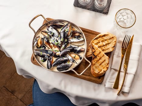 Delicious black mussels in creamy cheese and garlic sauce. Mediterranean black mussels in metal pan on wooden cutting board with grilled bread on restaurant table background, top view