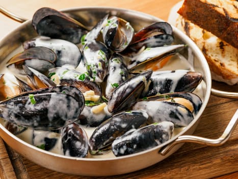 Delicious black mussels in creamy cheese and garlic sauce. Mediterranean black mussels in metal pan on wooden cutting board with grilled bread on wooden background