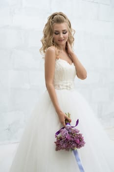 Gorgeous young bride in an exquisite dress posing with a violet bouquet in studio