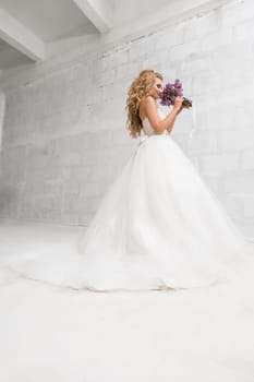 Young bride in beautiful white dress enjoying the aroma of her exquisite violet bouquet