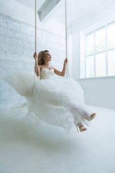 Gorgeous young bride in an exquisite dress posing on swing in white studio