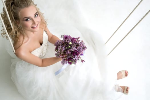 Gorgeous young bride posing in studio sitting on swing with an exquisite pink and violet bouquet